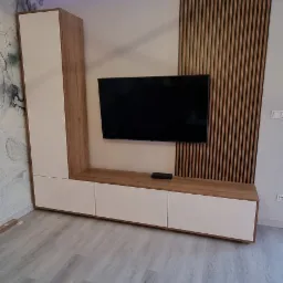 Nowoczesna szafka RTV na wymiar z białymi frontami i drewnianymi akcentami, podwieszony telewizor na tle drewnianych lameli, oświetlenie LED podsufitowe i designerska lampa wisząca.