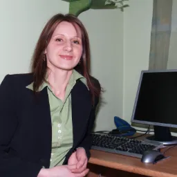 Kobieta w czarnej marynarce i jasnozielonej koszuli siedzi przy biurku z komputerem, klawiaturą i zszywaczem, uśmiechając się do kamery.