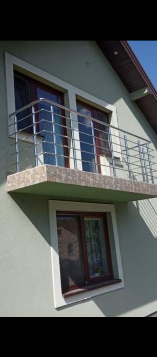 Balkon z balustradą ze stali nierdzewnej, wykończony płytkami imitującymi kamień, zamontowany przy oknie w domu o jasnej elewacji.