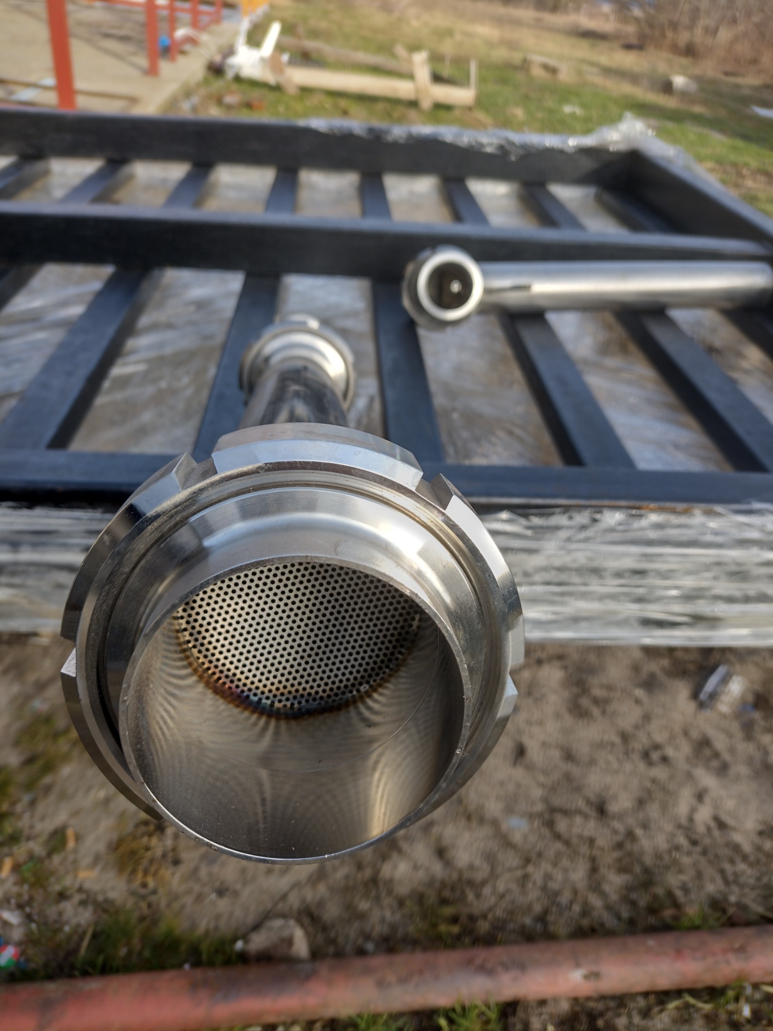 Elementy balustrady ze stali nierdzewnej z perforacją, przygotowane do montażu, leżące na czarnej konstrukcji kratowej.