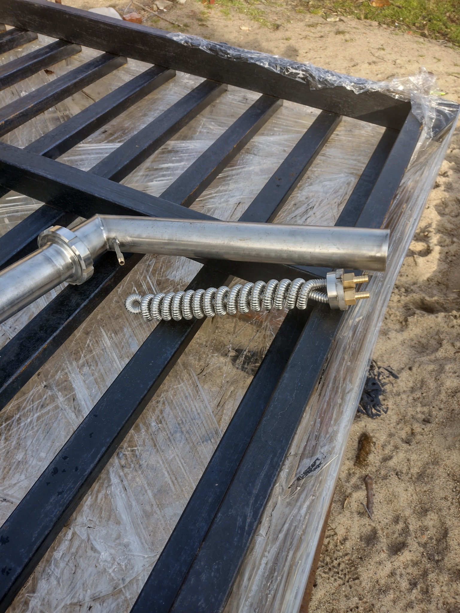 Elementy konstrukcji stalowej: rura ze stali nierdzewnej z końcówką kołnierzową i grzałka spiralna, ułożone na czarnej, zabezpieczonej folią ramie z profili stalowych.