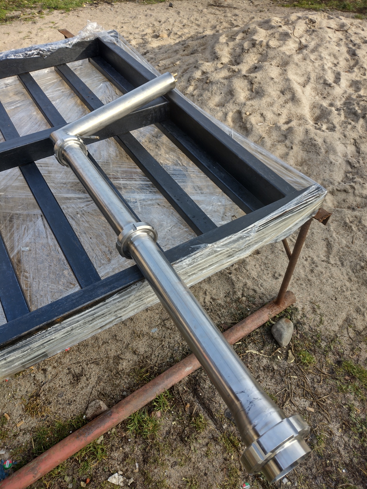 Element balustrady nierdzewnej z łącznikami, leżący na czarnej, metalowej konstrukcji zabezpieczonej folią stretch, na tle piaszczystego podłoża.