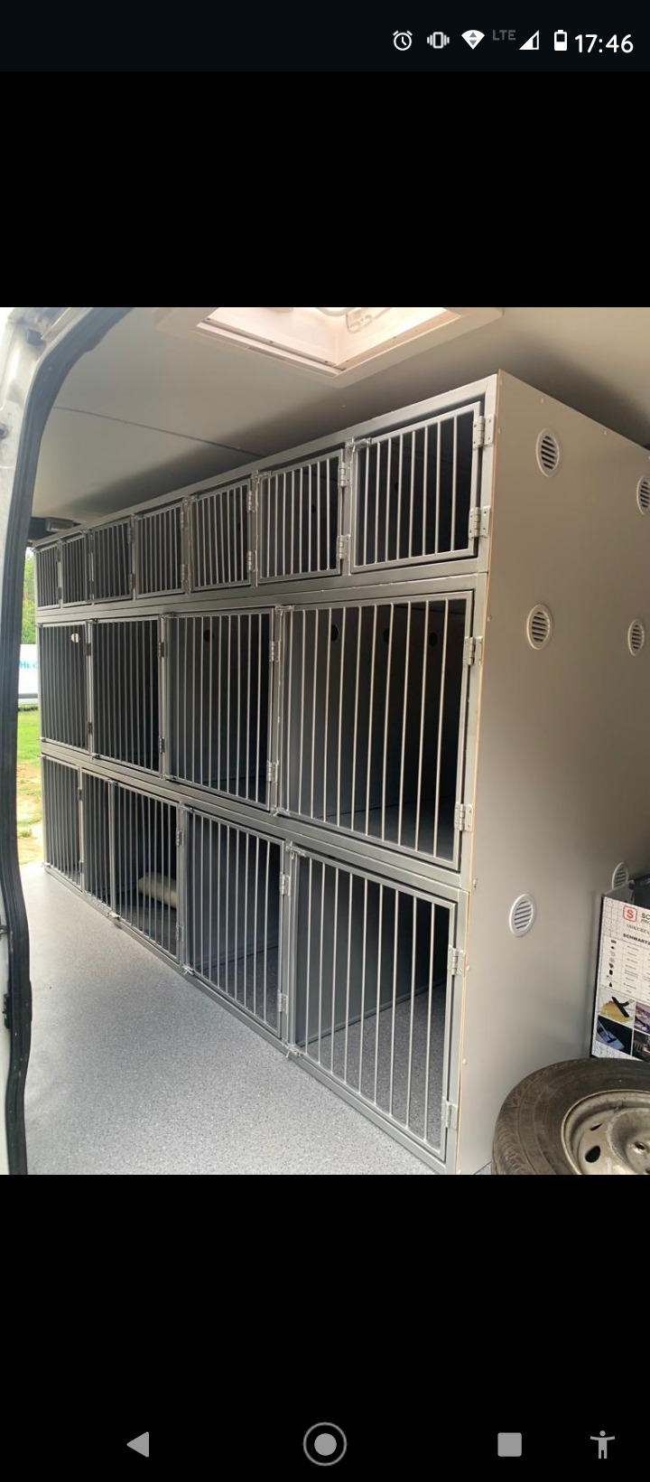 Wnętrze pojazdu z zamontowanymi klatkami z metalowymi prętami, ułożonymi w rzędach i kolumnach, szare ściany z otworami wentylacyjnymi, widoczna opona i ulotka informacyjna.