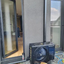 Czarna zewnętrzna jednostka pompy ciepła zamontowana na szarym żwirowym podłożu obok otwartych drzwi balkonowych, widoczny przewód odprowadzający skropliny, narzędzia i przewody serwisowe obok...