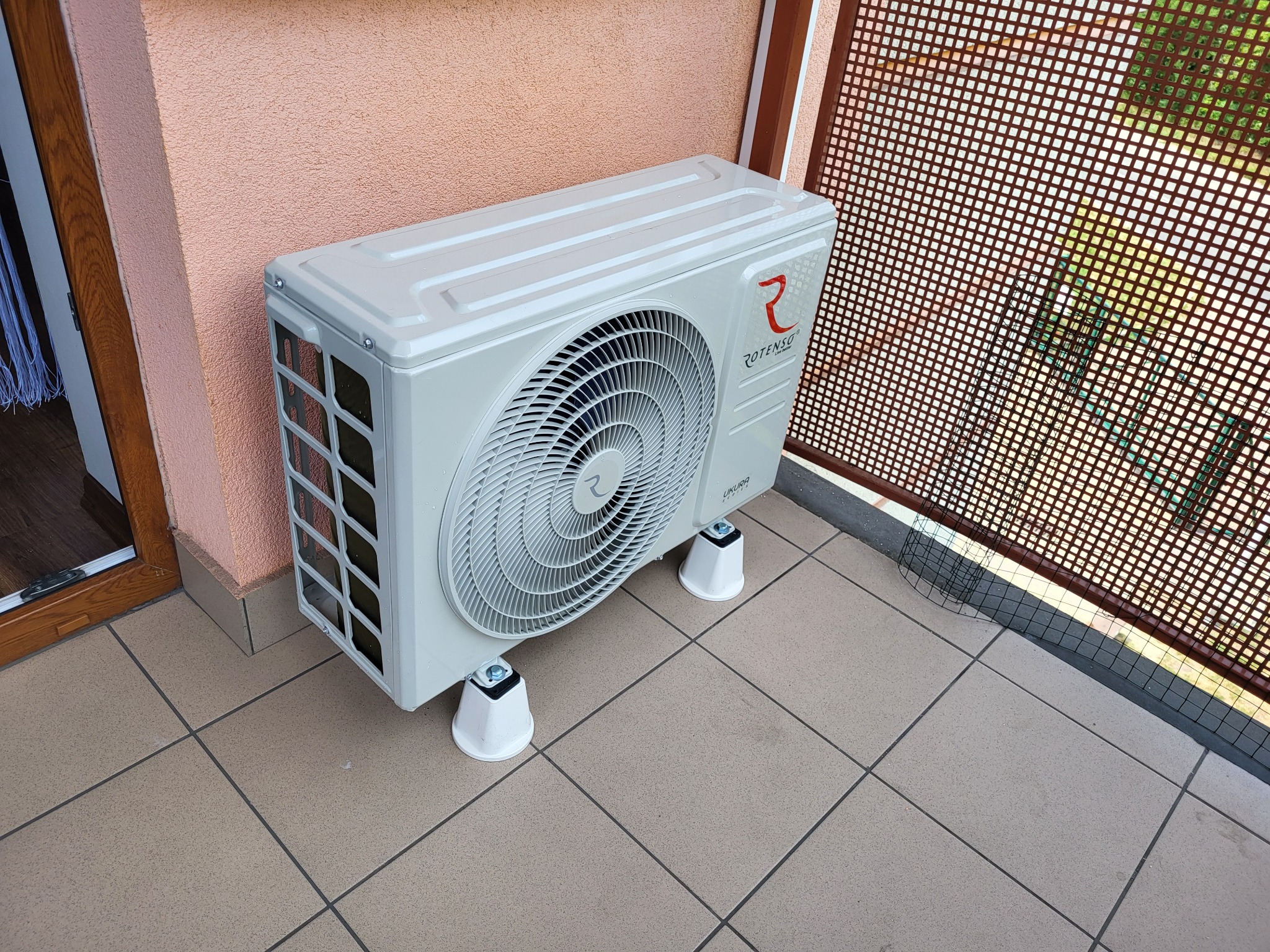Biała jednostka zewnętrzna klimatyzatora Rotenso Ukura zamontowana na balkonie na białych podkładkach antywibracyjnych.