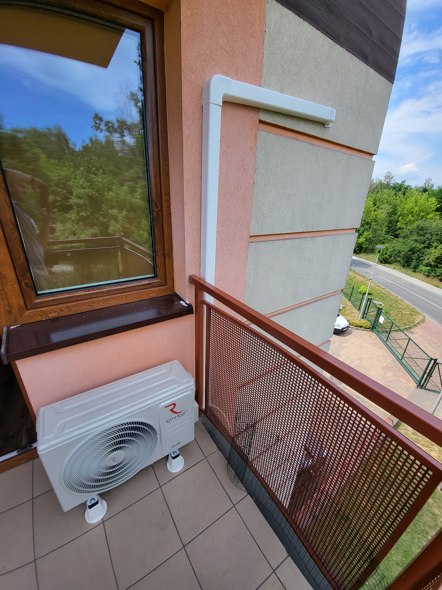 Biała jednostka zewnętrzna klimatyzacji Rotenso Ukura zamontowana na balkonie bloku z brązową balustradą, widok z góry na okolicę.