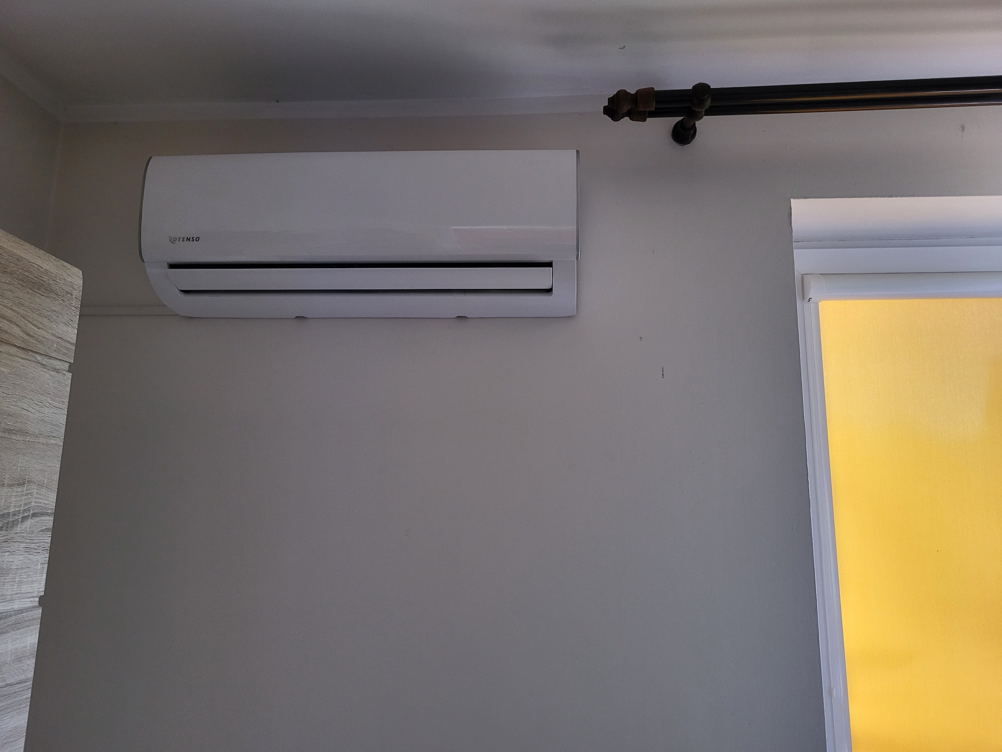 Biały klimatyzator marki Rotenso zamontowany na szarej ścianie, obok okna z żółtą szybą i czarną rurą dekoracyjną pod sufitem.