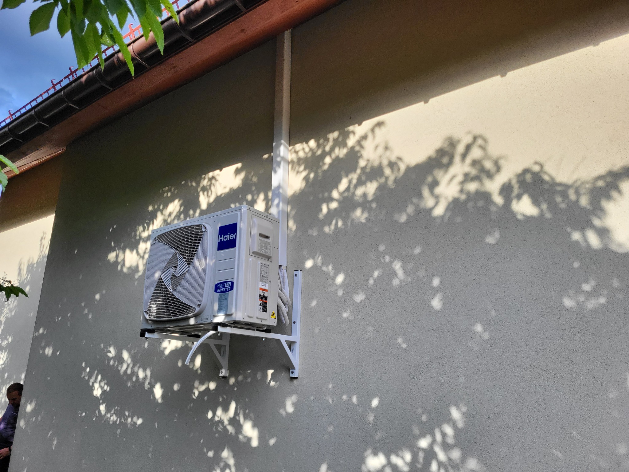 Zewnętrzna jednostka klimatyzatora Haier zamontowana na białym wsporniku na szarej ścianie budynku, z widocznymi cieniami liści i fragmentem dachu z rynną.