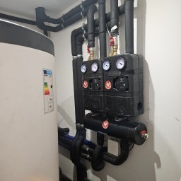 JN Hydro Usługi Instalacyjne - Zamknięty system grzewczy z czarnymi rurami w izolacji, manometrami i pompami obiegowymi, przylegający do białego zasobnika ciepłej wody z etykietą energetyczną.