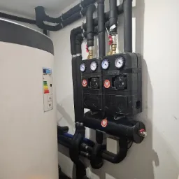 Zamknięty system grzewczy z czarnymi rurami w izolacji, manometrami i pompami obiegowymi, przylegający do białego zasobnika ciepłej wody z etykietą energetyczną.