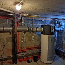 Montaż urządzenia grzewczego w piwnicy z widocznymi rurami, przewodami i narzędziami, oświetlone żarówką pod sufitem z surowego betonu.