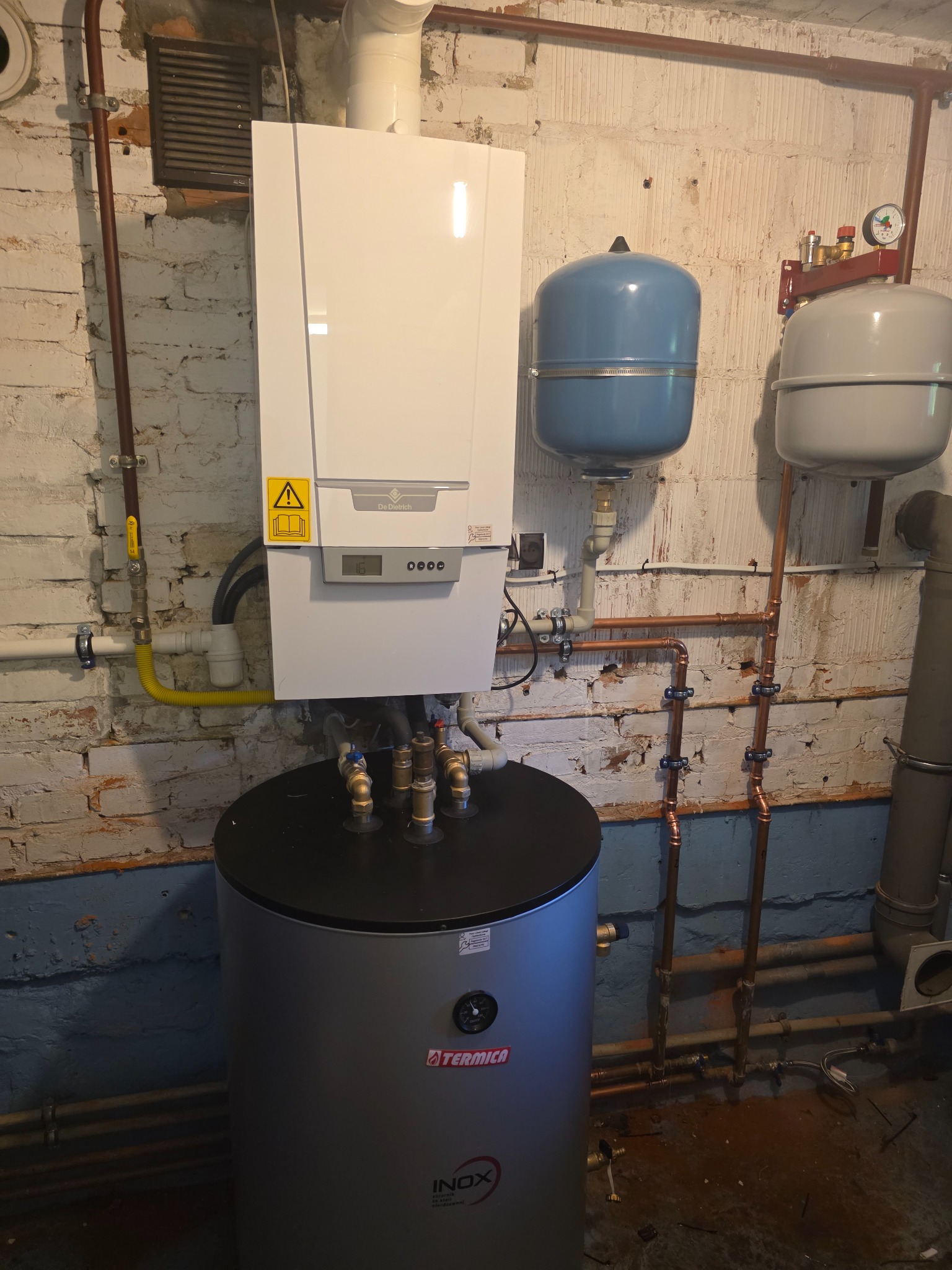 Nowoczesny piec gazowy De Dietrich z zasobnikiem Termica Inox i instalacją hydrauliczną na tle ściany z cegły w piwnicy.