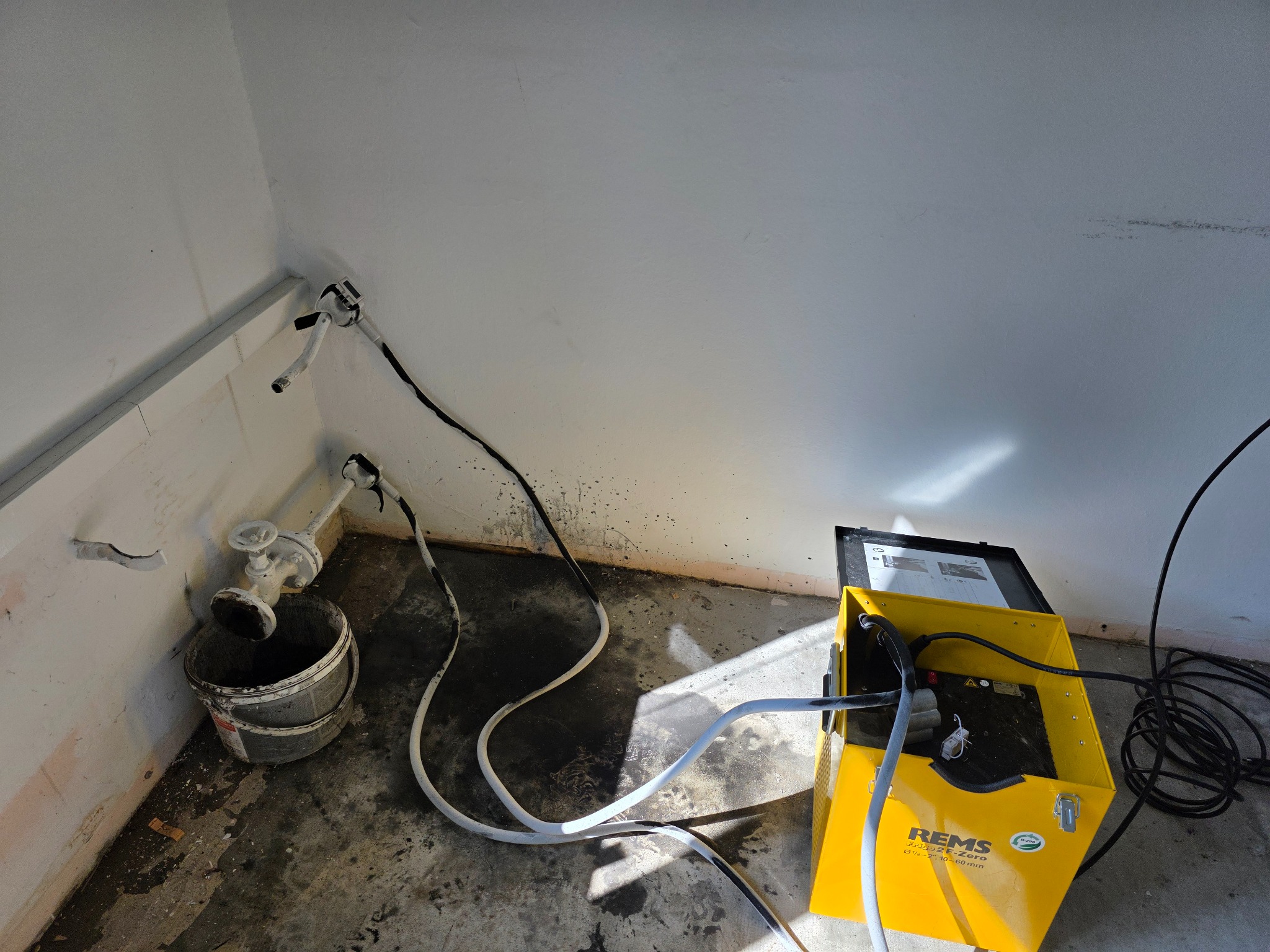 Żółta maszyna REMS do przepłukiwania instalacji grzewczej, podłączona do rur przy ścianie w rogu pomieszczenia. Widoczne węże i wiadro pod zaworem.