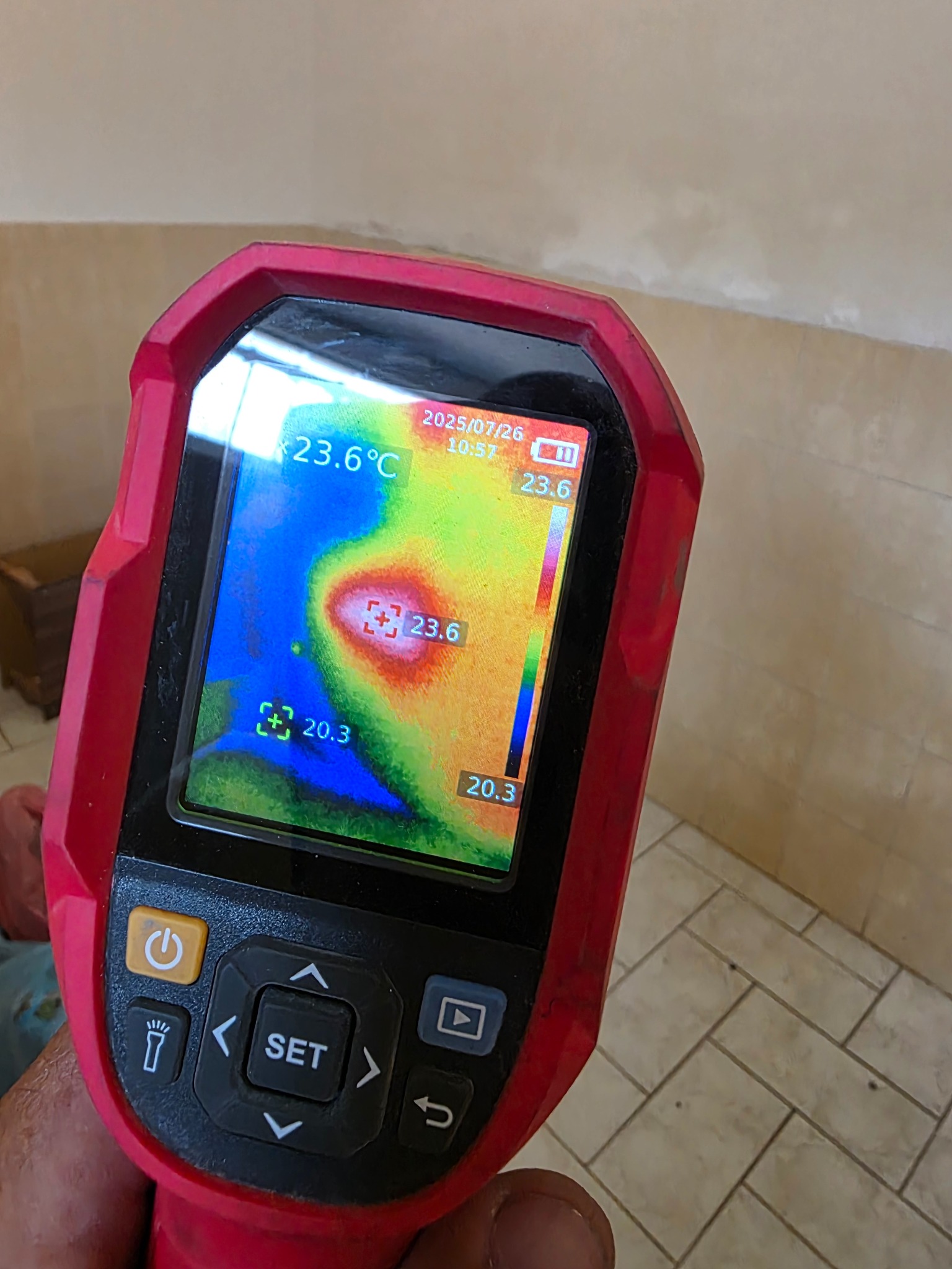 Termowizja ściany ukazuje rozkład temperatur na ekranie urządzenia, z datą 2025/07/26. Widoczna mozaika podłogowa i fragment ściany w tle.