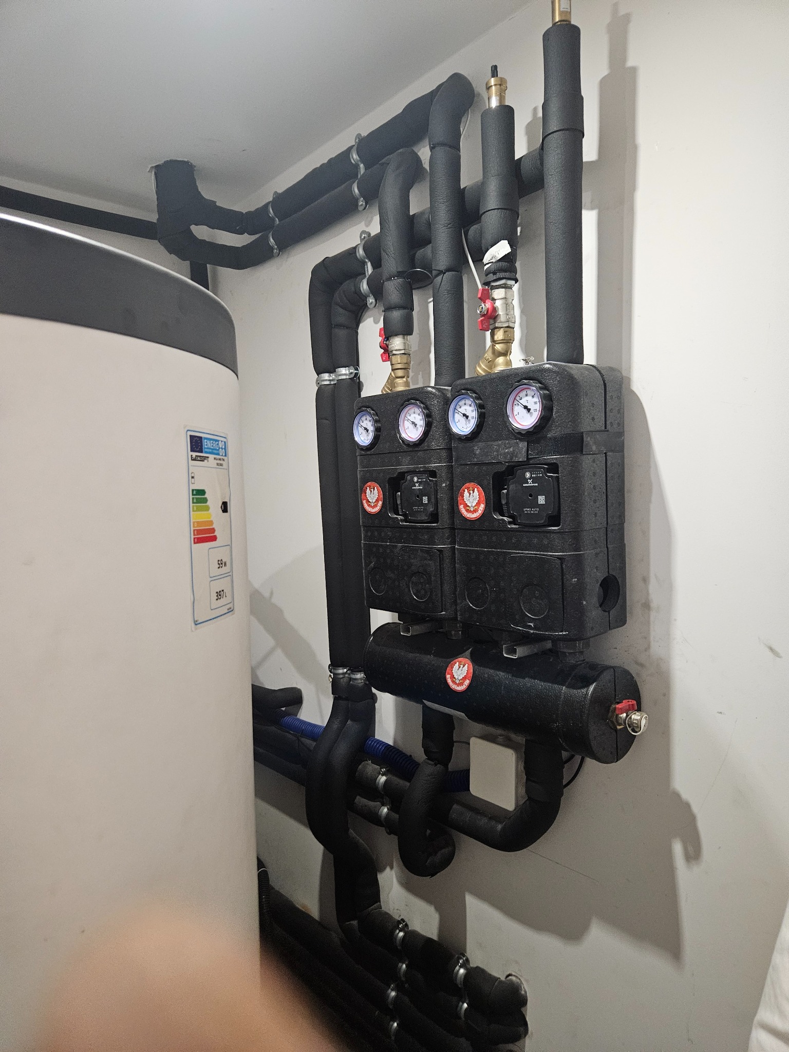 Zamknięty system grzewczy z czarnymi rurami w izolacji, manometrami i pompami obiegowymi, przylegający do białego zasobnika ciepłej wody z etykietą energetyczną.