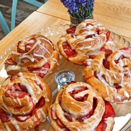 Canappka - Szklany talerz z pięcioma bułeczkami cynamonowymi ze świeżymi truskawkami i lukrem, obok wazon z lawendą, na drewnianym stole.