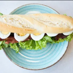 Kanapka bagietka z jajkiem na twardo, boczkiem i sałatą, ułożona na turkusowym talerzu w stylu rustykalnym, widok z góry.