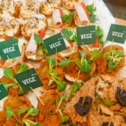 Różnorodne wegetariańskie kanapki na białej, ażurowej tacy, z elementami dekoracyjnymi takimi jak kiełki, szynka wegetariańska, warzywa i zielone flagi z napisem 'VEGE', na tle jasnych mebli...