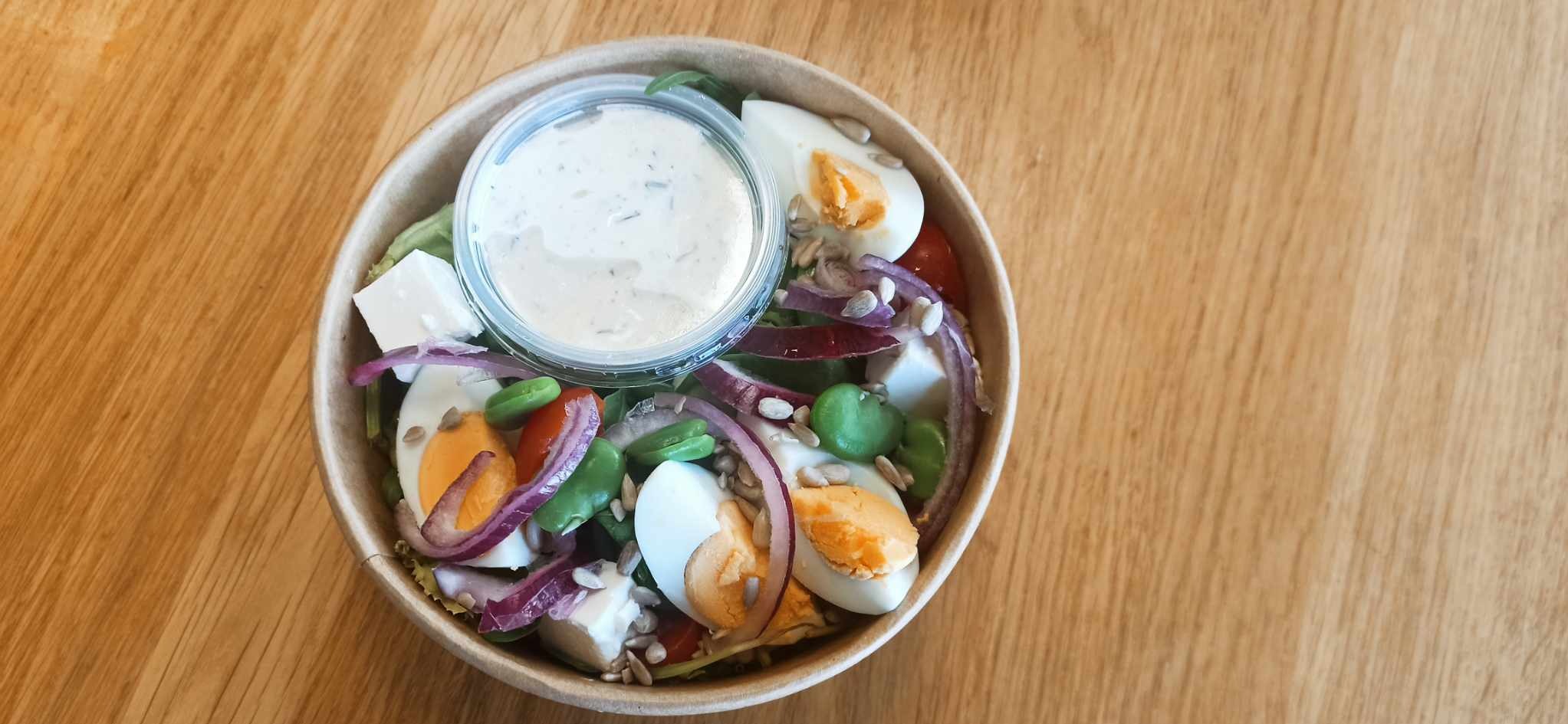 Sałatka w papierowej misce na drewnianym stole, zawierająca jajka na twardo, ser feta, czerwoną cebulę, zielone fasolki, pomidorki koktajlowe i nasiona słonecznika, z sosem w plastikowym pojemniku.