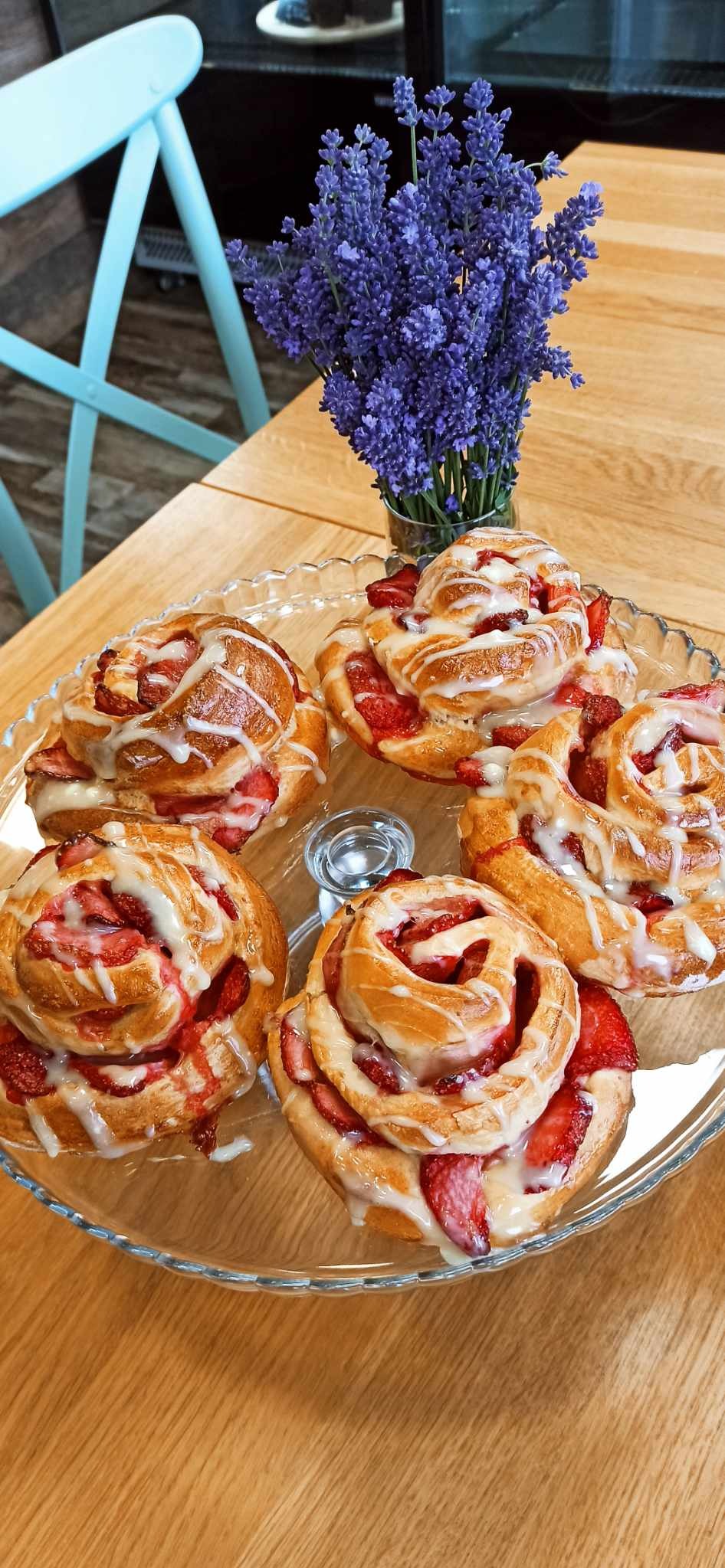 Szklany talerz z pięcioma bułeczkami cynamonowymi ze świeżymi truskawkami i lukrem, obok wazon z lawendą, na drewnianym stole.