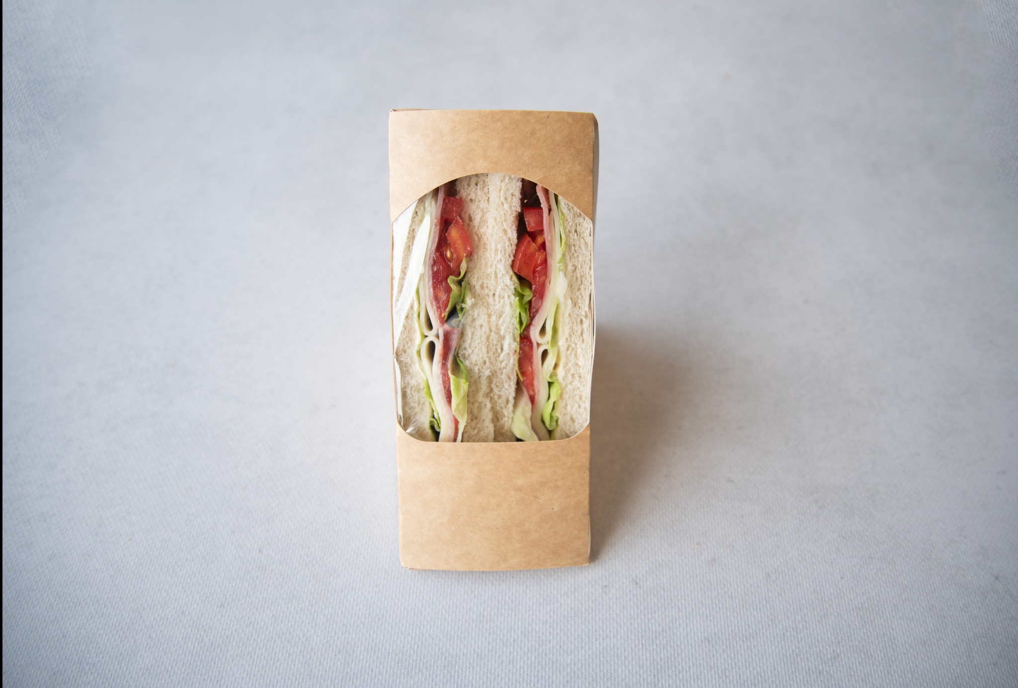 Kanapka przekrojona na pół, z widocznymi warstwami sałaty, pomidora i szynki, umieszczona w brązowym papierowym opakowaniu z okienkiem, na jasnoszarym tle.
