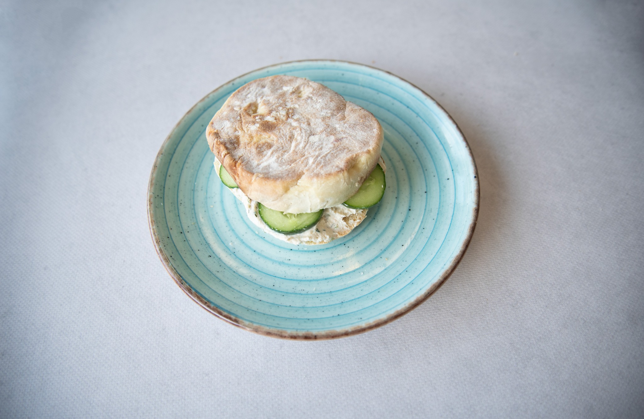 Kanapka z ogórkiem i kremowym serem na turkusowym talerzu z widoczną fakturą, ułożona na białym obrusie, zrobiona w jasnym świetle.