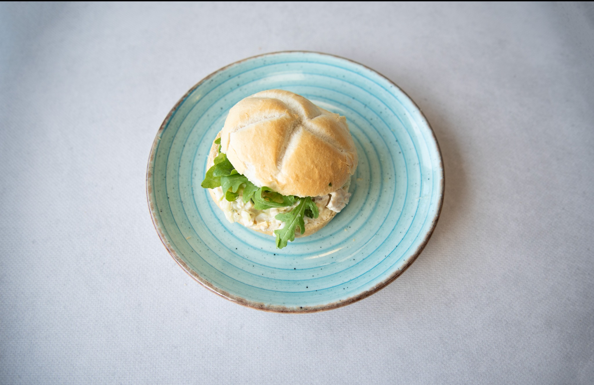 Kanapka z jasnego pieczywa, kurczakiem i rukolą na turkusowym, ceramicznym talerzu z brązową obwódką.