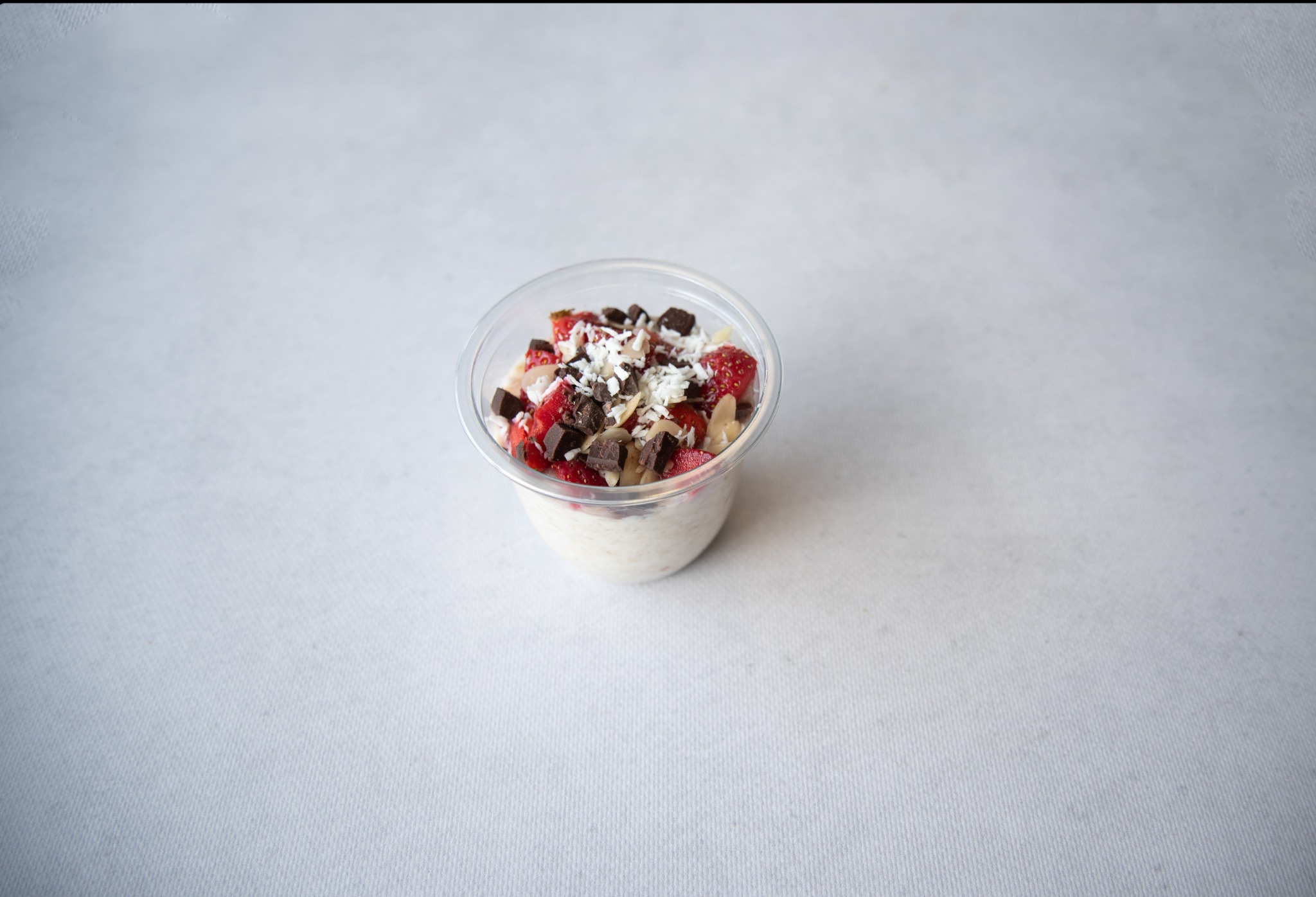 Deser w przezroczystym plastikowym kubku: ryż na mleku z pokrojonymi truskawkami, kawałkami czekolady i wiórkami kokosowymi, na jasnym tle.