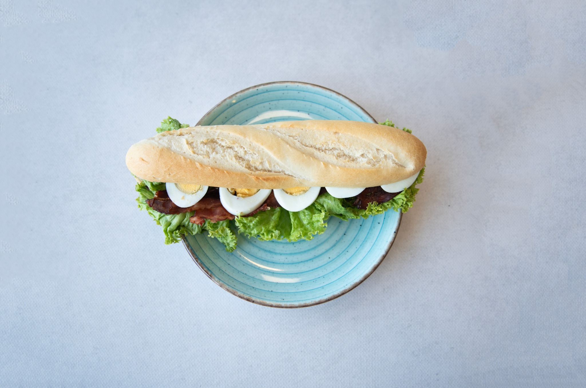 Kanapka z bagietki z jajkiem na twardo, boczkiem i sałatą, ułożona na turkusowym talerzu z efektem spękań, widok z góry.