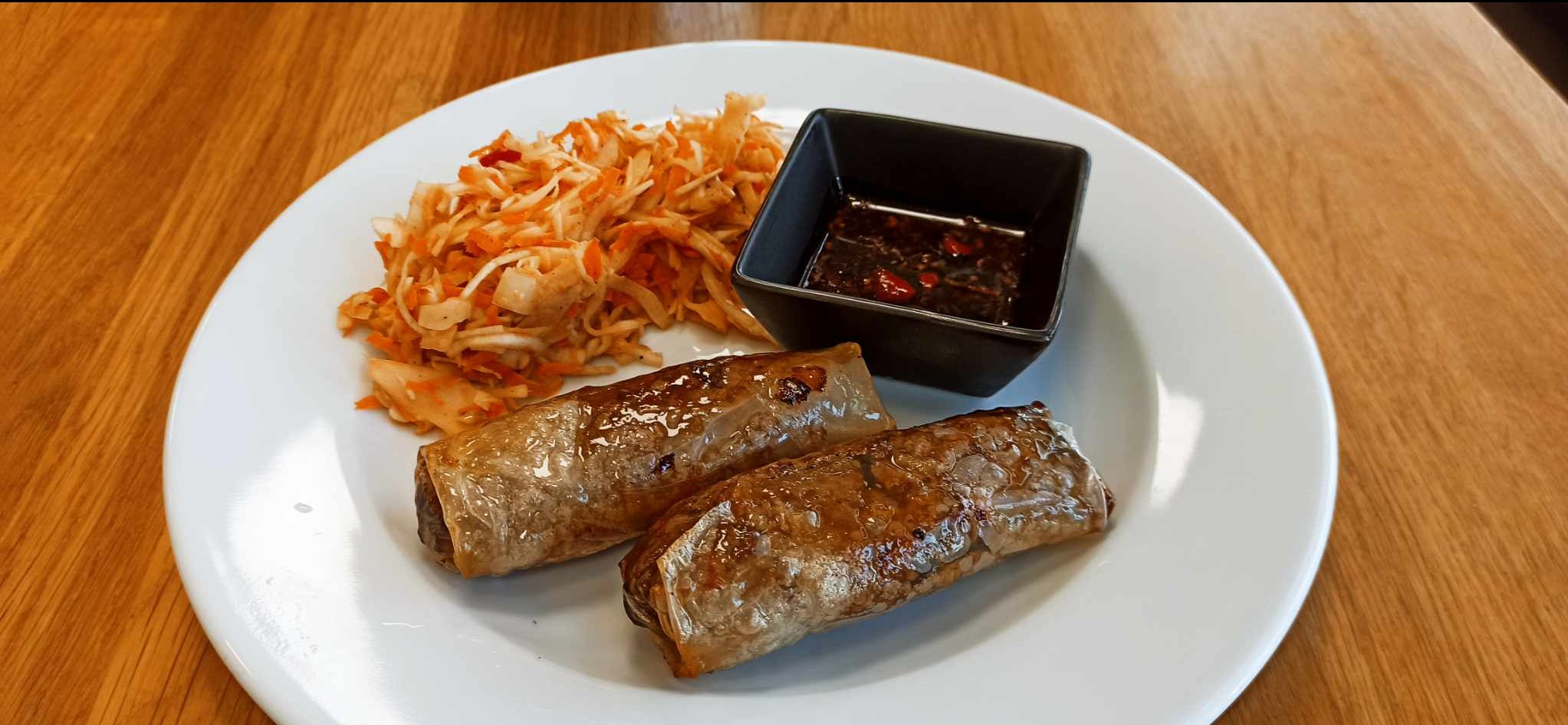 Dwa smażone sajgonki na białym talerzu, podane z surówką z marchewki i kapusty oraz sosem chili w czarnym kwadratowym naczyniu.