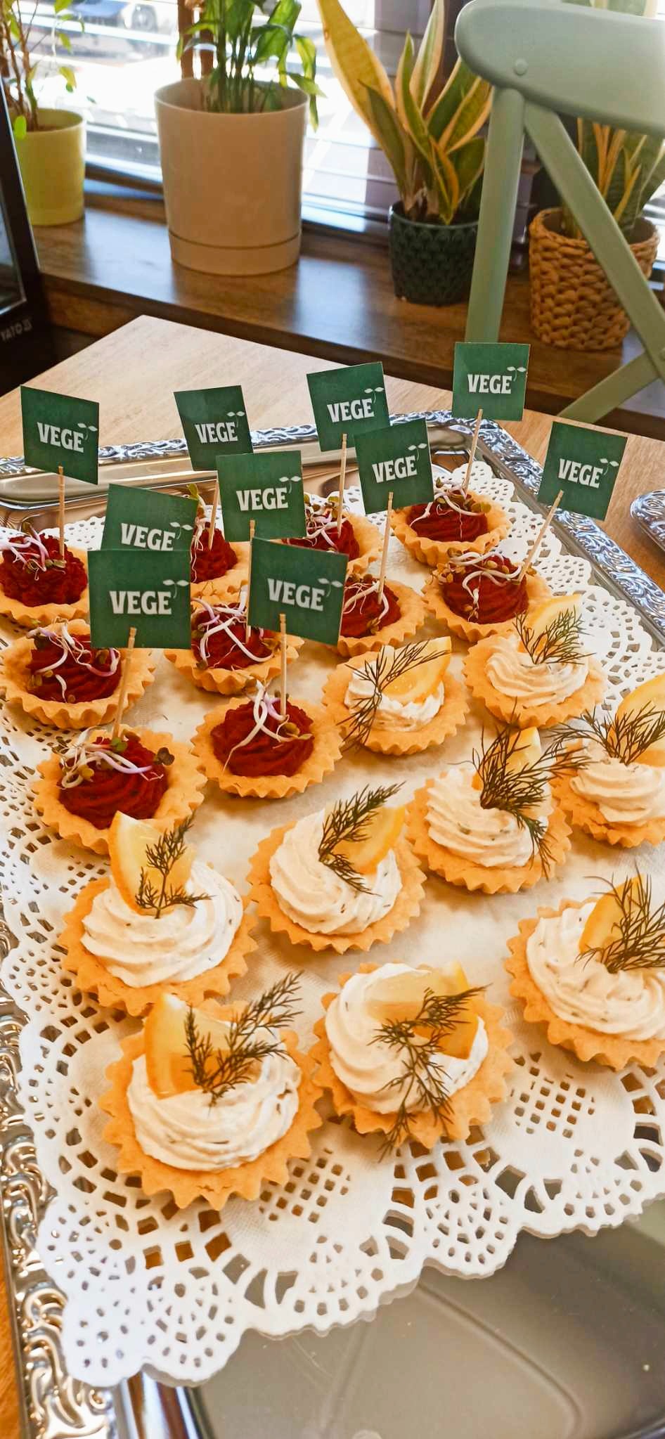 Talerz z tartaletkami: część z kremem i cytryną z koperkiem, część z buraczkową pastą i kiełkami, wszystkie z zielonymi chorągiewkami 'VEGE', na srebrnej tacy z serwetką.