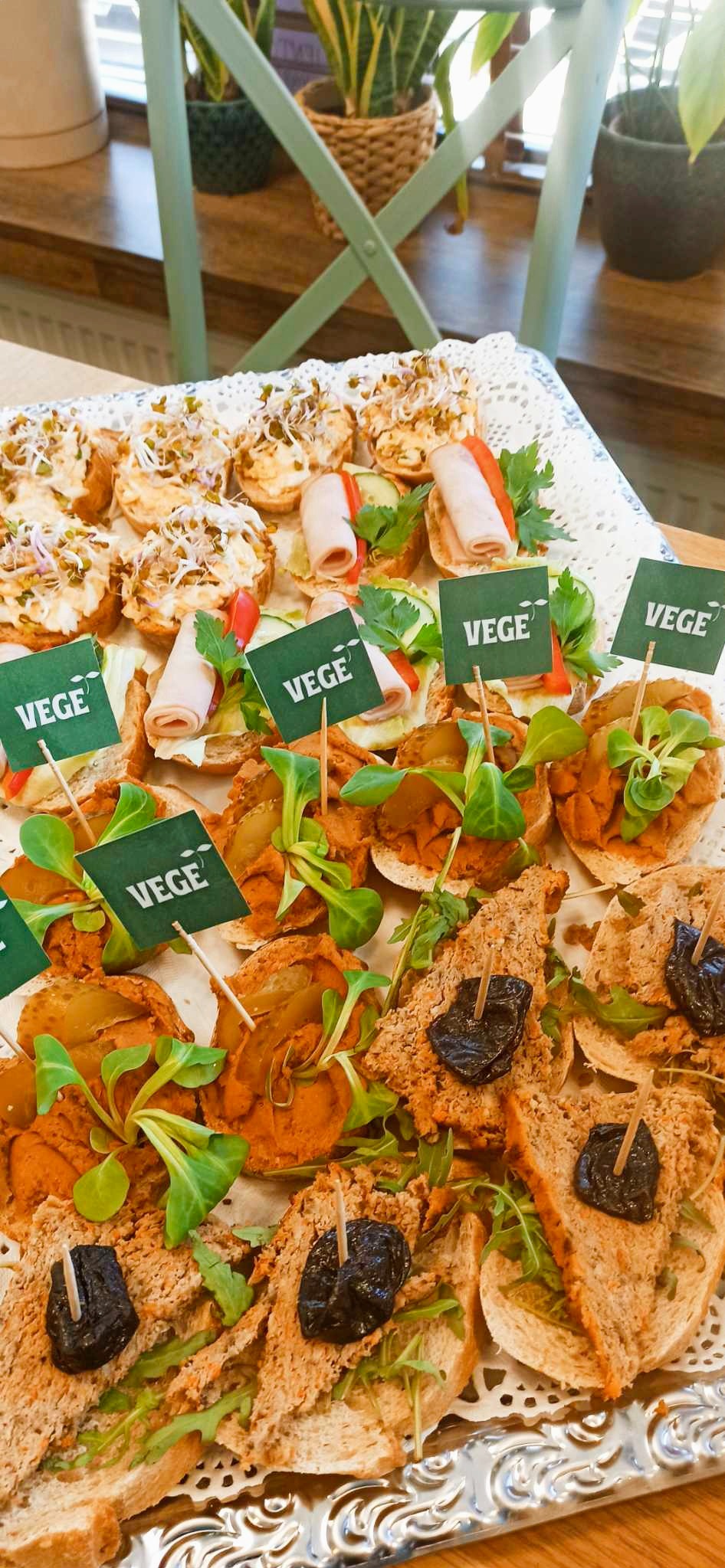 Różnorodne wegetariańskie kanapki na białej, ażurowej tacy, z elementami dekoracyjnymi takimi jak kiełki, szynka wegetariańska, warzywa i zielone flagi z napisem 'VEGE', na tle jasnych mebli...