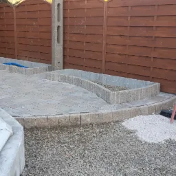 Układanie kostki brukowej wokół basenu i rabat kwiatowych, widoczne narzędzia i materiały budowlane, brązowe ogrodzenie w tle.