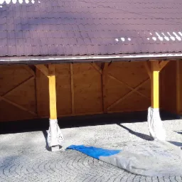 Drewniana konstrukcja z brązowym dachem krytym dachówką, częściowo osłonięta folią ochronną, na wybrukowanym podłożu, z wózkiem transportowym w tle.