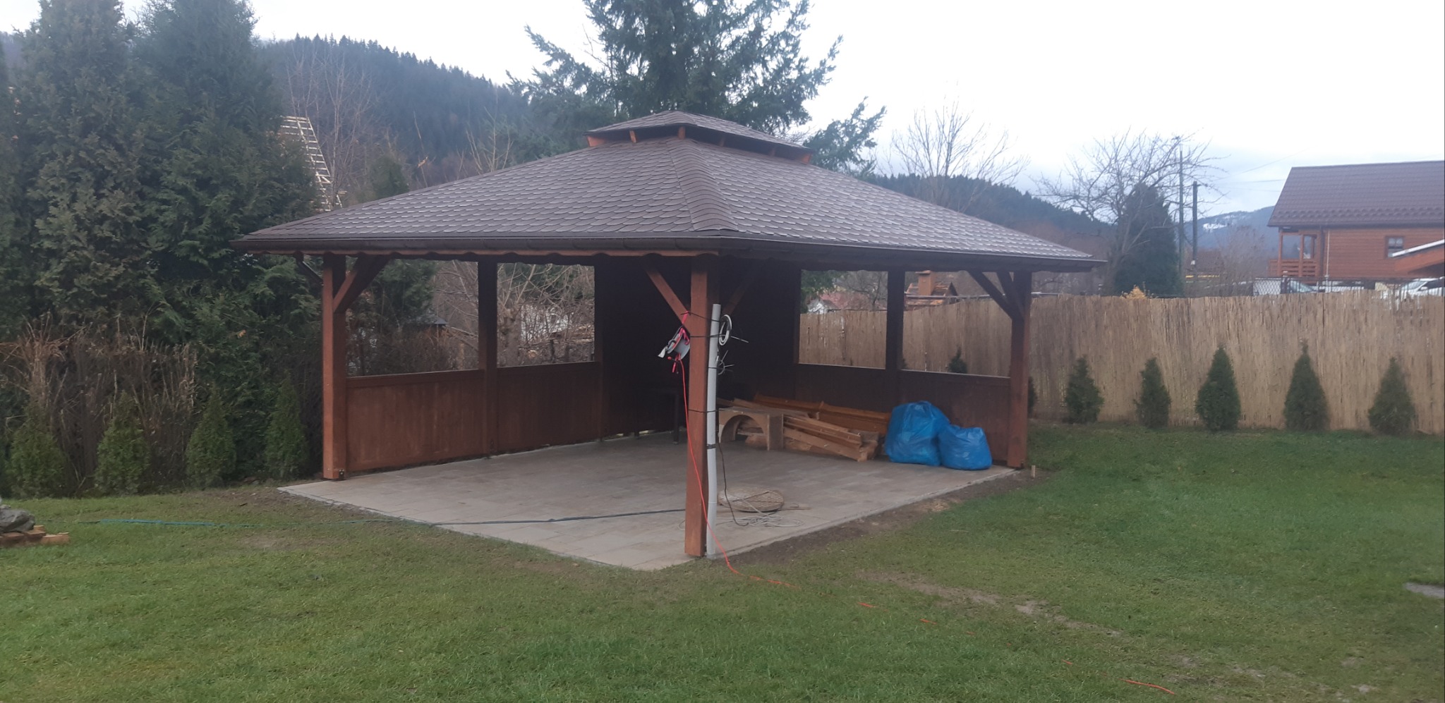 Drewniana altana z brązowym dachem, częściowo zabudowana, na betonowej podłodze. Wewnątrz niezabezpieczone drewniane elementy i worki. W tle zielony trawnik, ogrodzenie z trzciny i zalesione wzgórza.