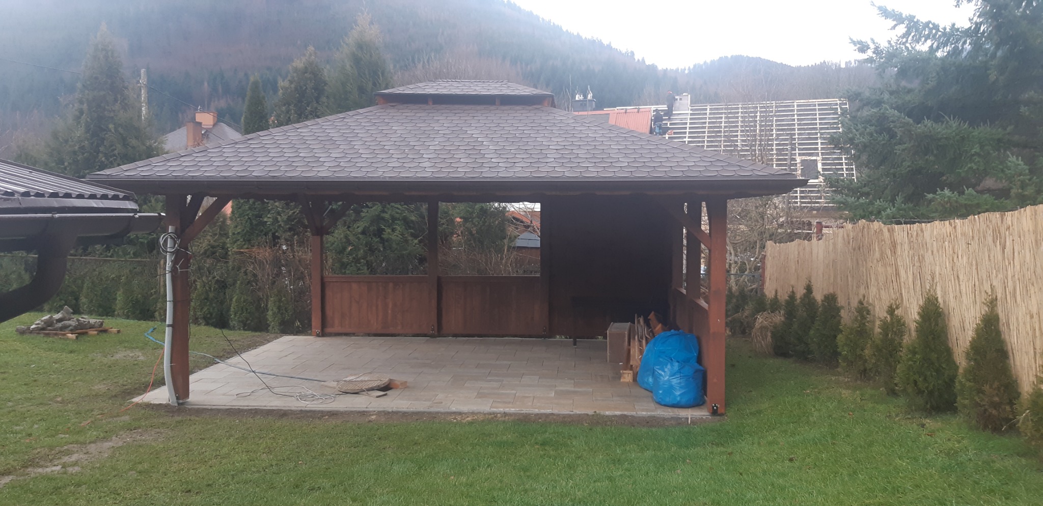 Drewniana altana ogrodowa z brązowym dachem z gontów bitumicznych i częściowym obłożeniem ścianek, na wyłożonym kostką brukową patio, widok na zalesione wzgórze w tle.