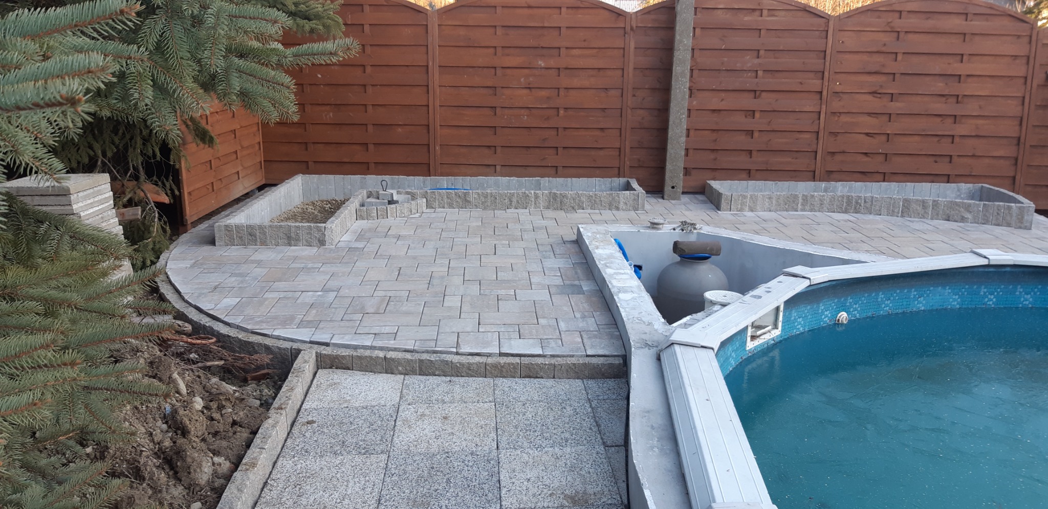 Ułożona kostka brukowa wokół basenu ogrodowego z drewnianym ogrodzeniem w tle i fragmentami zieleni