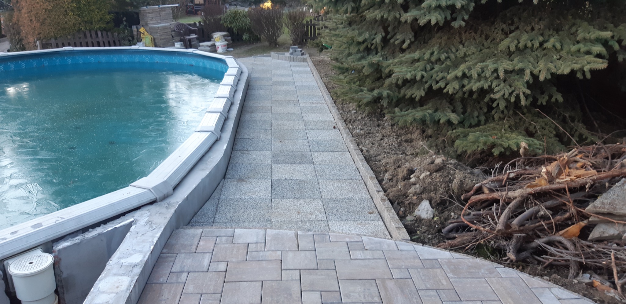 Świeżo ułożona ścieżka z kostki brukowej prowadząca do basenu ogrodowego, z widocznymi obrzeżami i przygotowanym terenem pod nasadzenia, w tle iglaki.