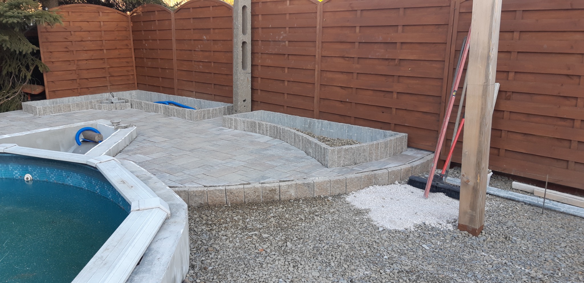 Układanie kostki brukowej wokół basenu i rabat kwiatowych, widoczne narzędzia i materiały budowlane, brązowe ogrodzenie w tle.