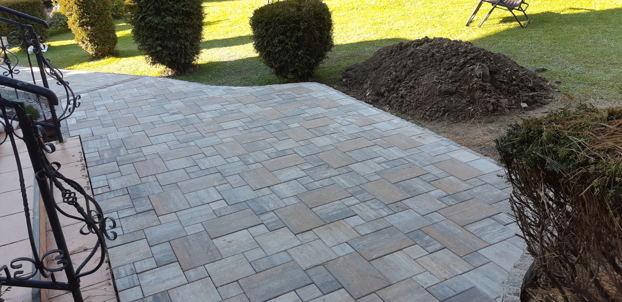 Ułożona kostka brukowa w odcieniach szarości i beżu, przylegająca do trawnika z krzewami i stertą ziemi, widoczna czarna, ozdobna balustrada schodów.