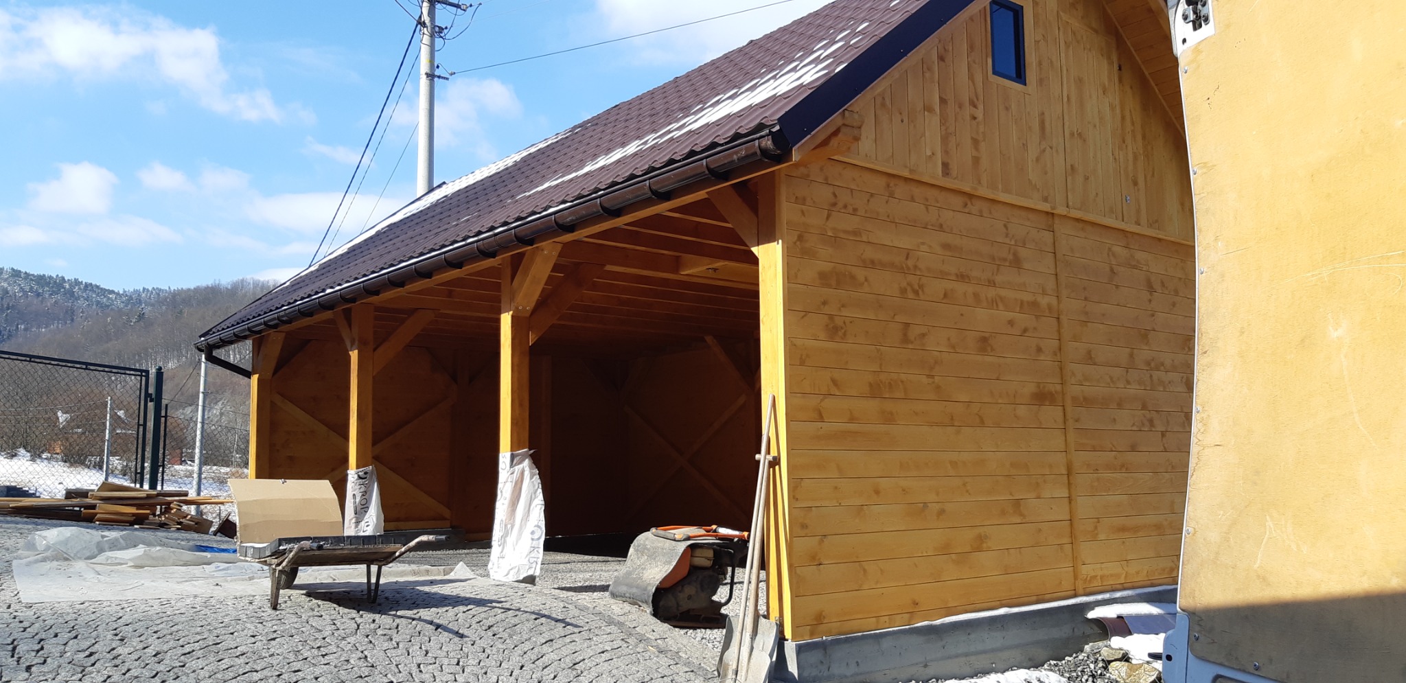 Nowo wybudowana, drewniana altana z brązowym dachem na posesji z kostki brukowej, widoczne narzędzia i materiały budowlane, w tle zalesione wzgórza.