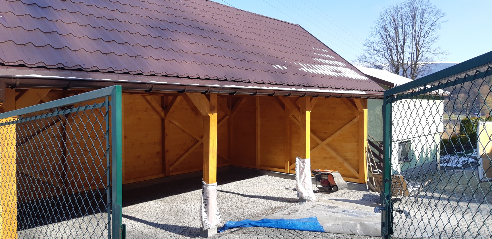 Nowo wybudowana, otwarta, drewniana konstrukcja z brązowym dachem z blachodachówki, widoczna zza zielonej, otwartej bramy wjazdowej, z elementami zabezpieczającymi słupy przed uszkodzeniem...