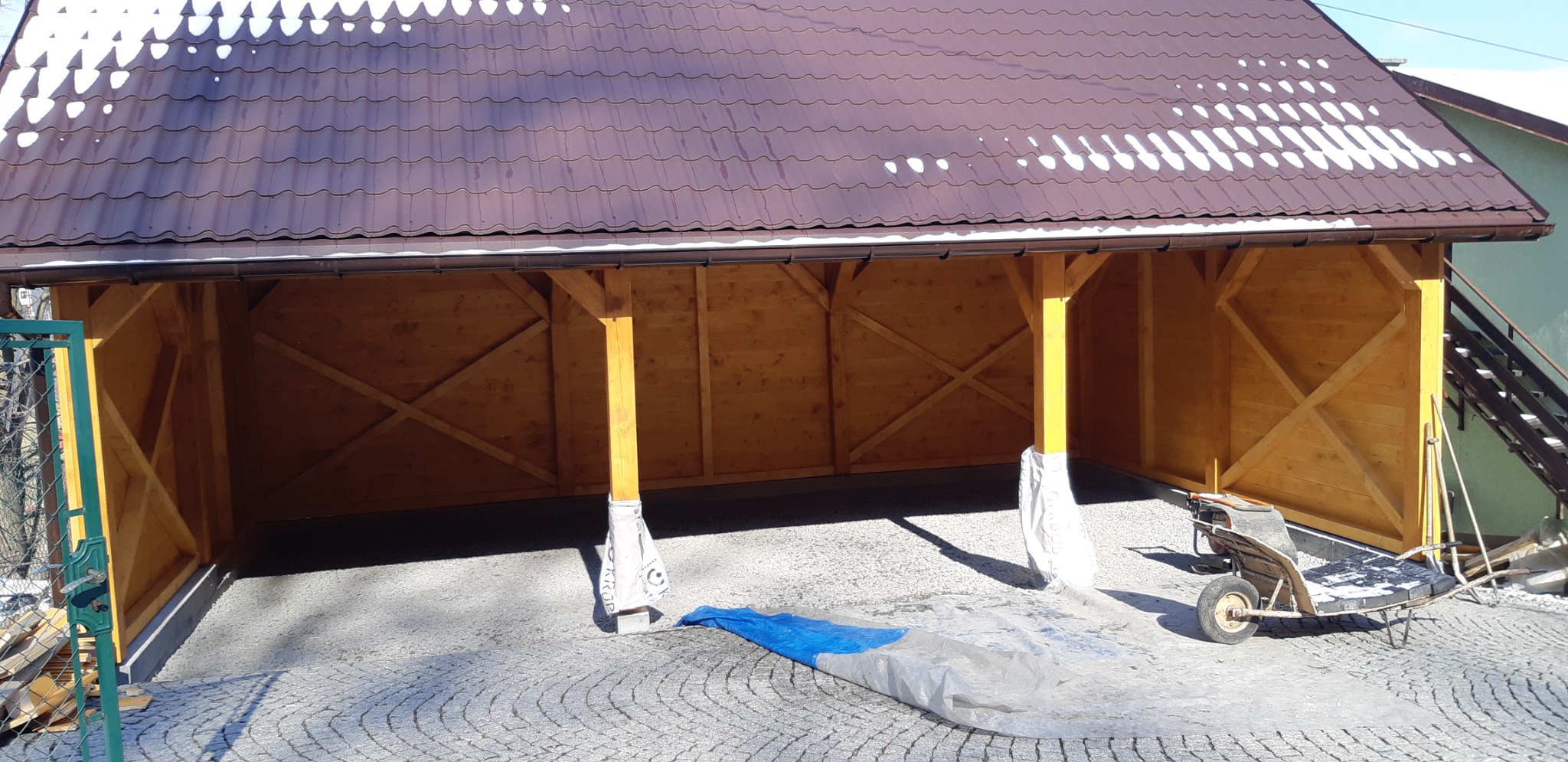 Drewniana konstrukcja z brązowym dachem krytym dachówką, częściowo osłonięta folią ochronną, na wybrukowanym podłożu, z wózkiem transportowym w tle.