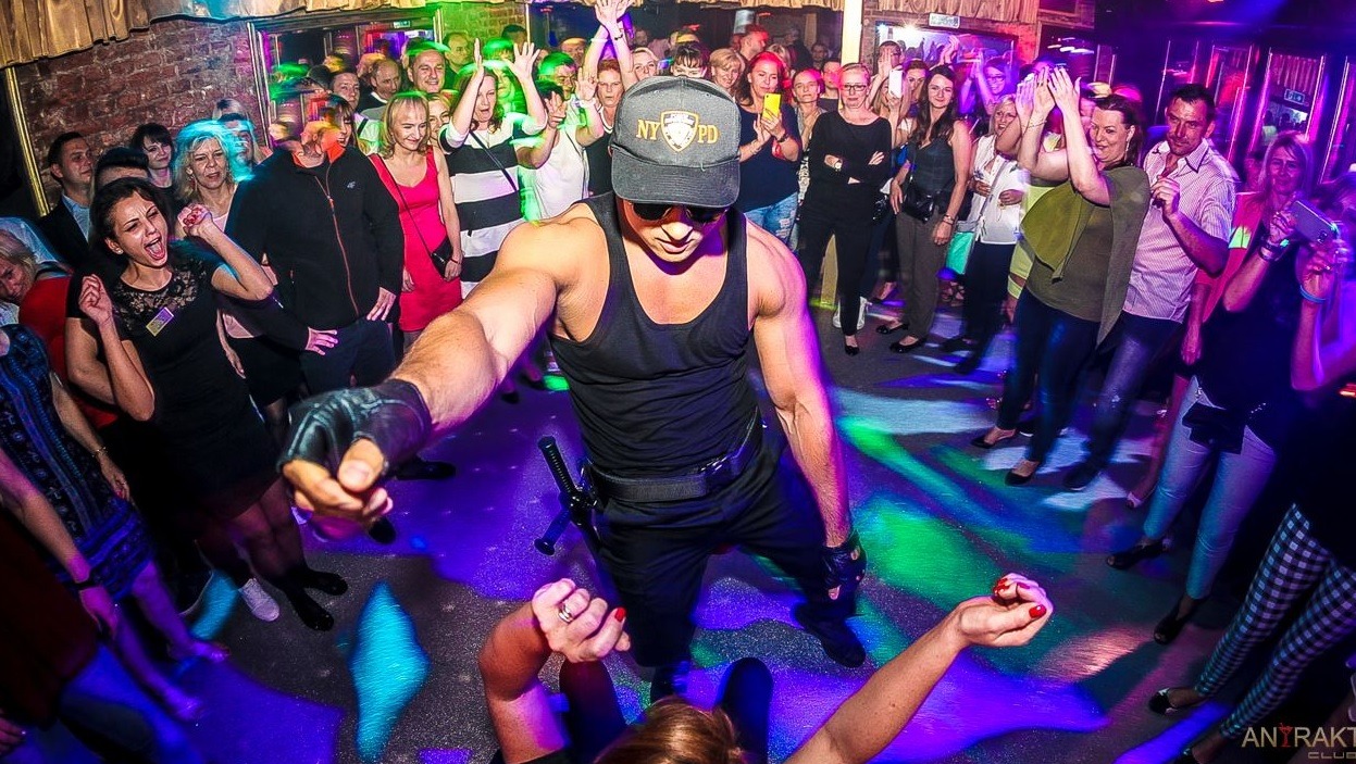 Mężczyzna w czarnym stroju i czapce NYPD w centrum uwagi na parkiecie, otoczony rozbawionym tłumem w klubie z kolorowymi światłami.