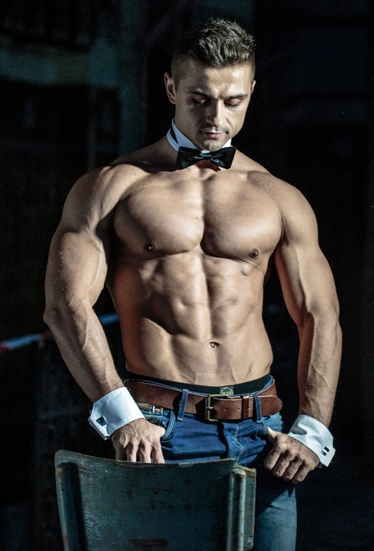 Mężczyzna o muskularnej sylwetce, ubrany w muszkę i mankiety koszuli, opiera ręce na oparciu krzesła, w tle ciemne otoczenie.