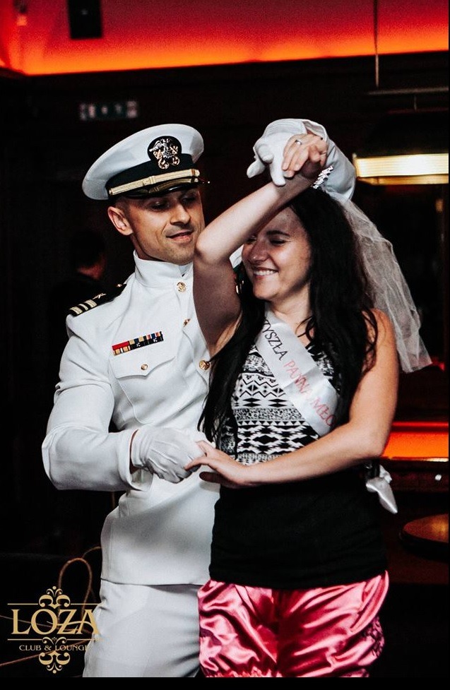 Mężczyzna w białym mundurze oficerskim tańczy z uśmiechniętą kobietą w welonie i szarfie 'Przyszła Panna Młoda' w ciemnym klubie z czerwonym oświetleniem.