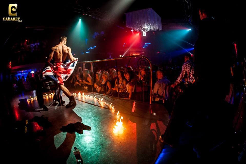 Mężczyzna bez koszuli, owinięty flagą Wielkiej Brytanii, stoi na scenie oświetlonej kolorowymi światłami, z płonącymi świecami i publicznością w tle.