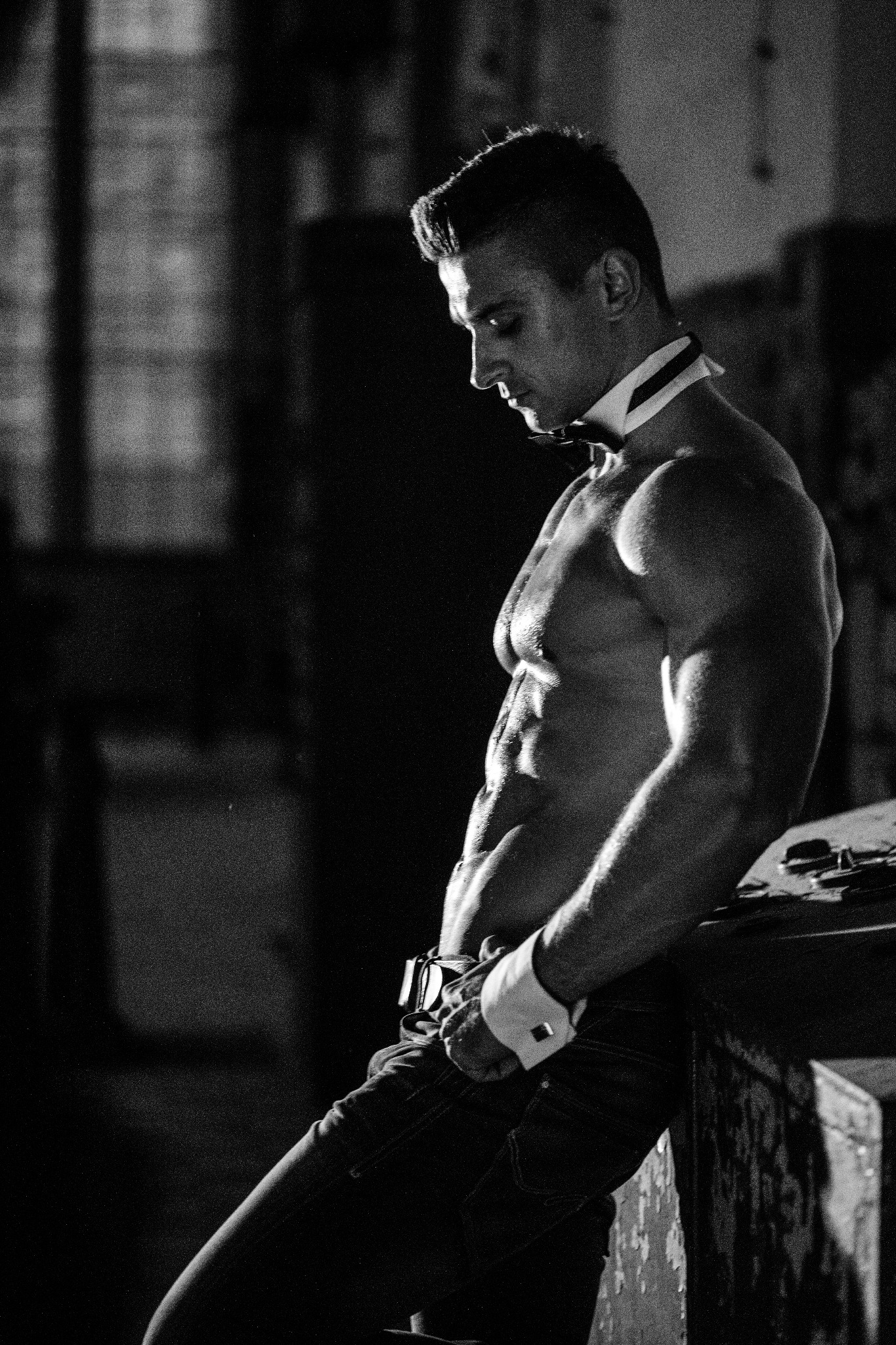 Czarno-białe zdjęcie muskularnego mężczyzny w jeansach i z muszką, opierającego się o stół w ciemnym pomieszczeniu, prawdopodobnie podczas występu.