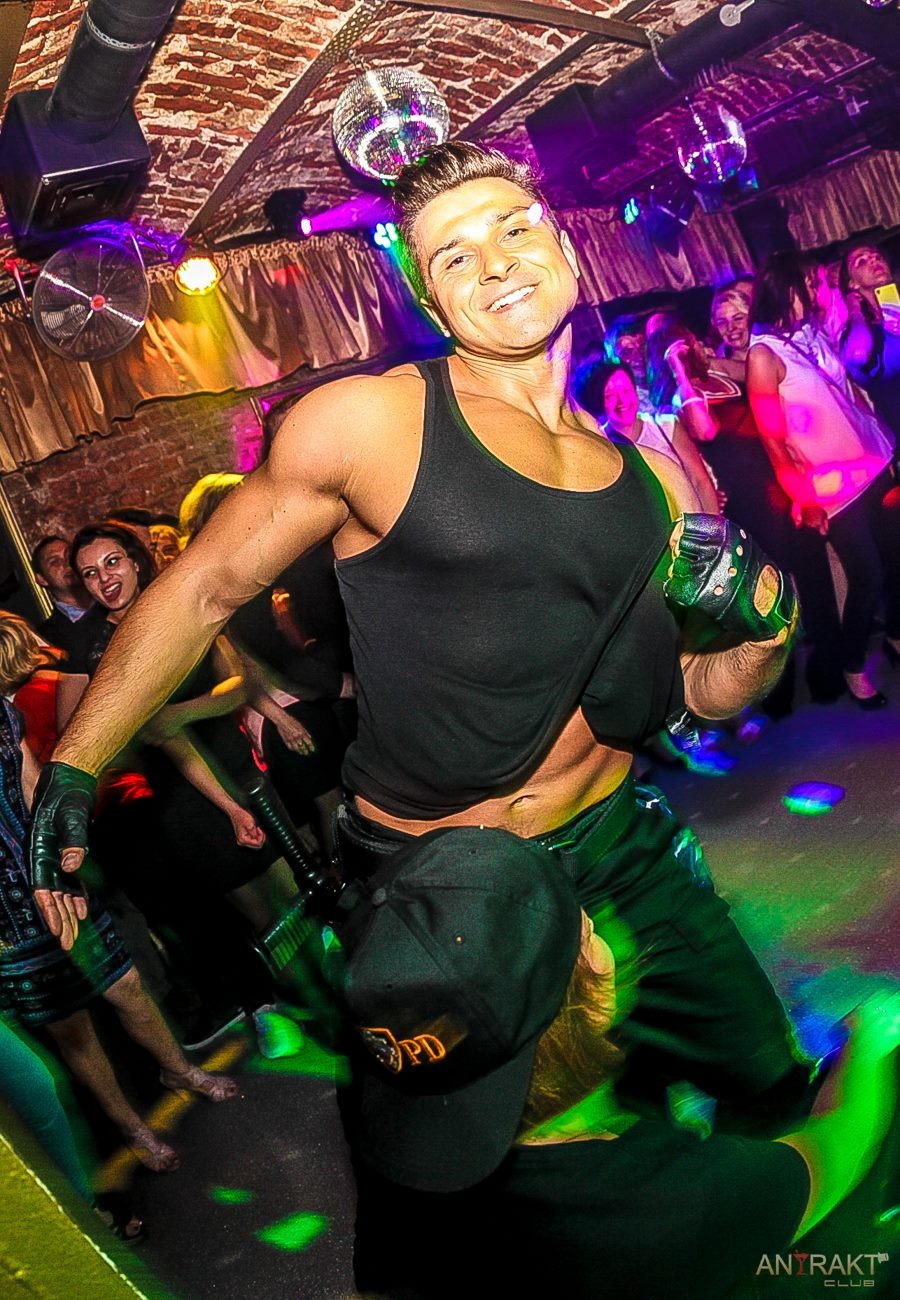 Mężczyzna w czarnym tank topie i skórzanych rękawicach tańczy w klubie z publicznością w tle, oświetlony kolorowymi światłami dyskotekowymi i z odblaskową kulą na suficie.