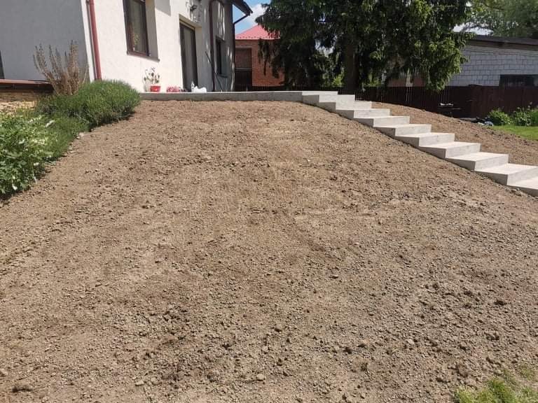 Przygotowany teren pod trawnik z betonowymi schodami prowadzącymi do domu. Ziemia wyrównana i spulchniona, gotowa do siewu trawy. Widoczne rośliny ozdobne przy ścianie budynku.